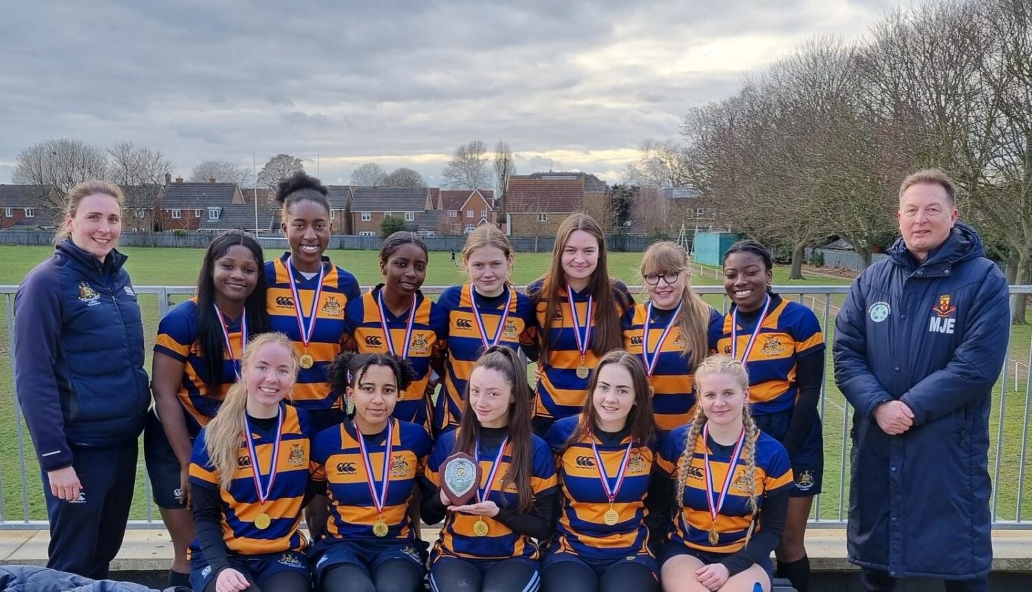 Participants in a Girls Rugby Tournament are seen posing together for a group photo, alongside their coaches, wearing gold medals around their necks.