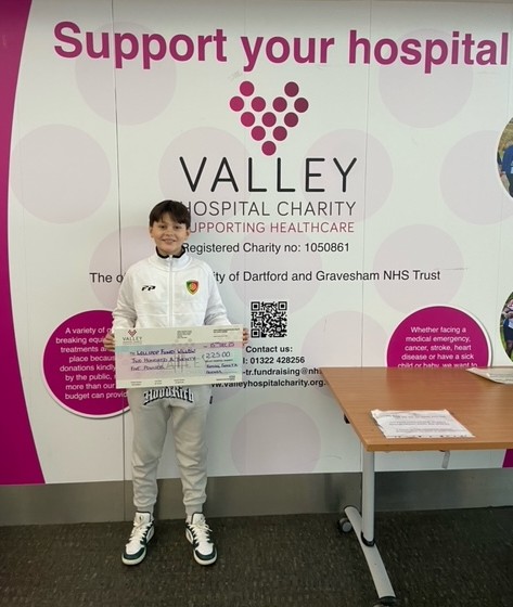 A boy with a large cheque for £225 is pictured in front of a 'support your hospital' sign at Darenth Valley hospital